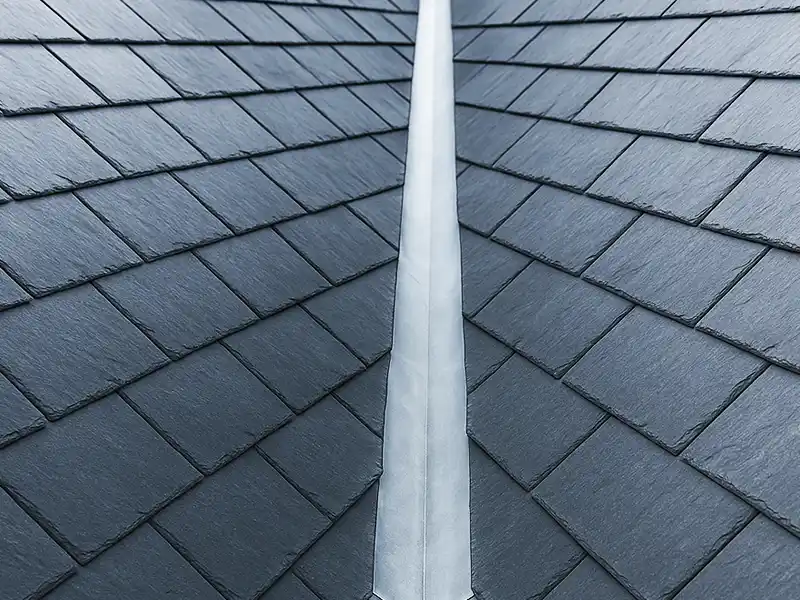 Gros plan d'une toiture en tuiles d'ardoise avec solin de noue métallique, montrant l'étanchéité noue toiture, où deux pans de toiture se rejoignent, et des rangées soignées de tuiles gris foncé qui se chevauchent.