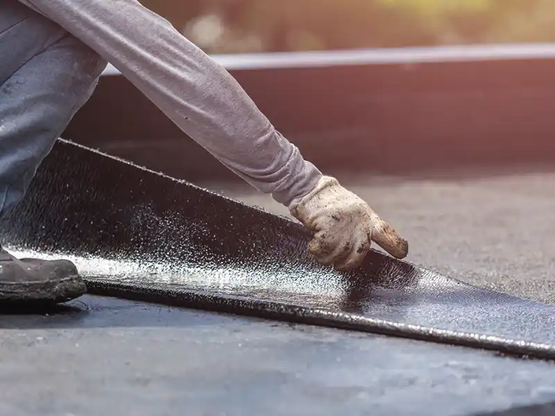 Une personne portant des gants et des manches longues installe une membrane d'étanchéité noire et adhésive sur un toit plat, en la lissant soigneusement à la main pour assurer une parfaite étanchéité toit terrasse.