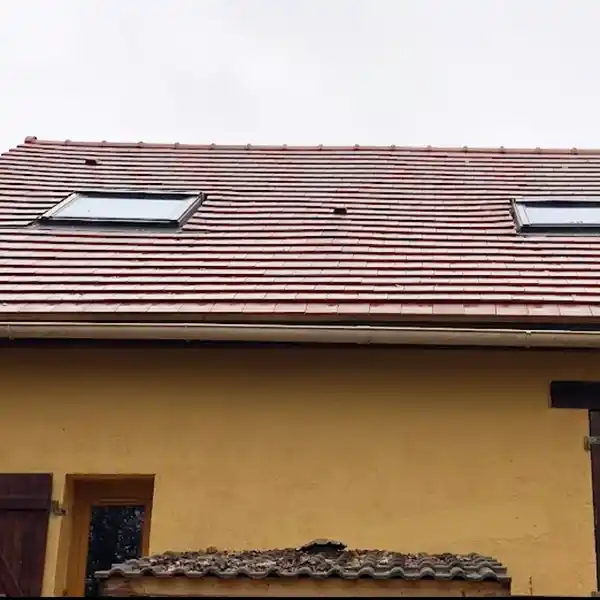 Une maison jaune au toit de tuiles rouges - remplacement de la toiture Loiret - après - est dotée de deux fenêtres à lucarne. Au-dessus, le ciel gris et couvert se profile, tandis qu'en dessous, il y a une petite structure couverte et un volet en bois sombre sur une fenêtre.
