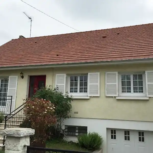 Une maison jaune clair avec un toit en tuiles rouges illustrant le remplacement de toiture (apr&egrave;s travaux), des volets blancs et une porte d'entr&eacute;e rouge. Des escaliers m&egrave;nent &agrave; l'entr&eacute;e et une porte de garage blanche se trouve en dessous. Des buissons et des plantes poussent pr&egrave;s des marches.