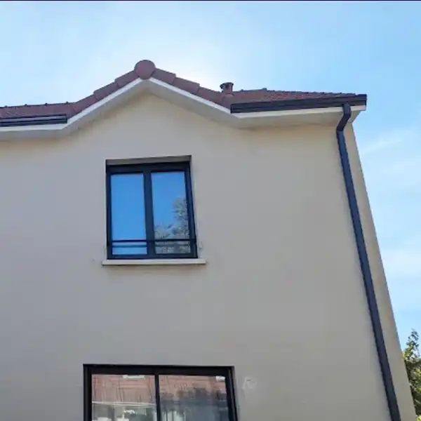 Une maison beige à deux étages avec une fenêtre à cadre sombre à l'étage supérieur et une fenêtre plus grande en dessous, avec un toit incliné en tuiles rouges et une gouttière grise - une inspiration idéale pour un Remplacement des gouttières dans le Loiret, sur fond de ciel bleu clair.